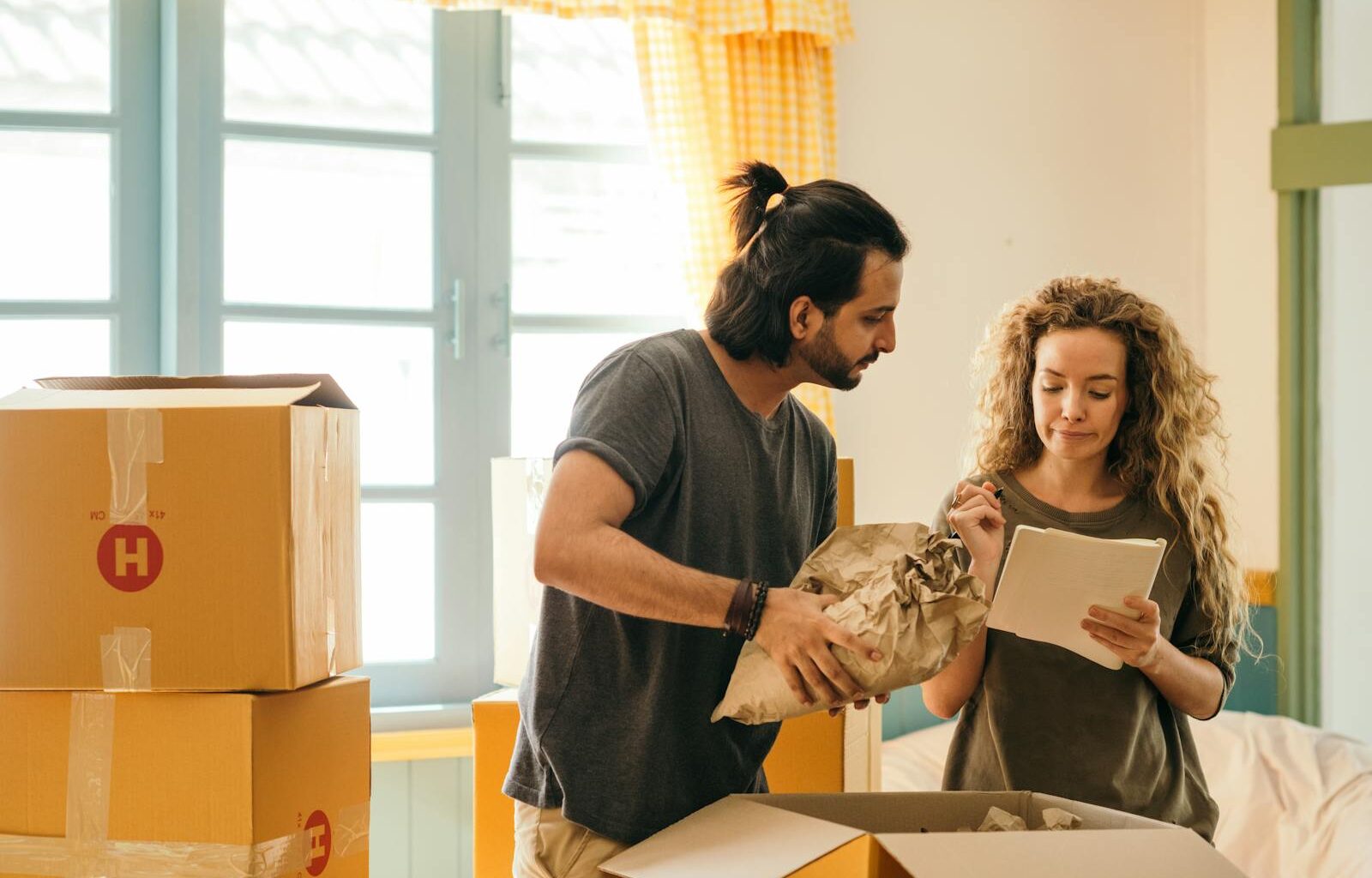 Cheerful young man and woman smiling while unpacking carton boxes with belongings in new apartment during relocation and looking at paper