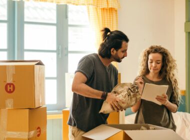 Cheerful young man and woman smiling while unpacking carton boxes with belongings in new apartment during relocation and looking at paper